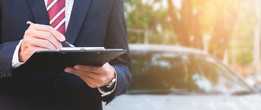 Man in suit looking at papers in board with a car in the background.