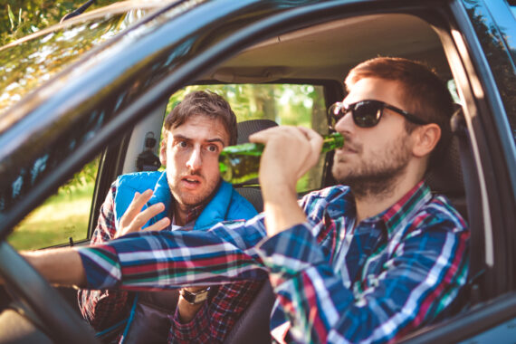 Two men in a car, driver is drinking a beer and passenger is objecting