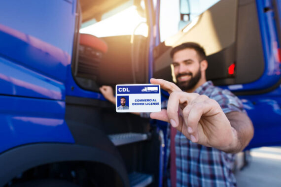 A smiling trucker stands outside of his truck showing his CDL license.